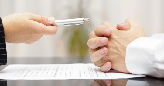A hand holding a pen across a paper on a desk and another person's hands folded on top of the paper.